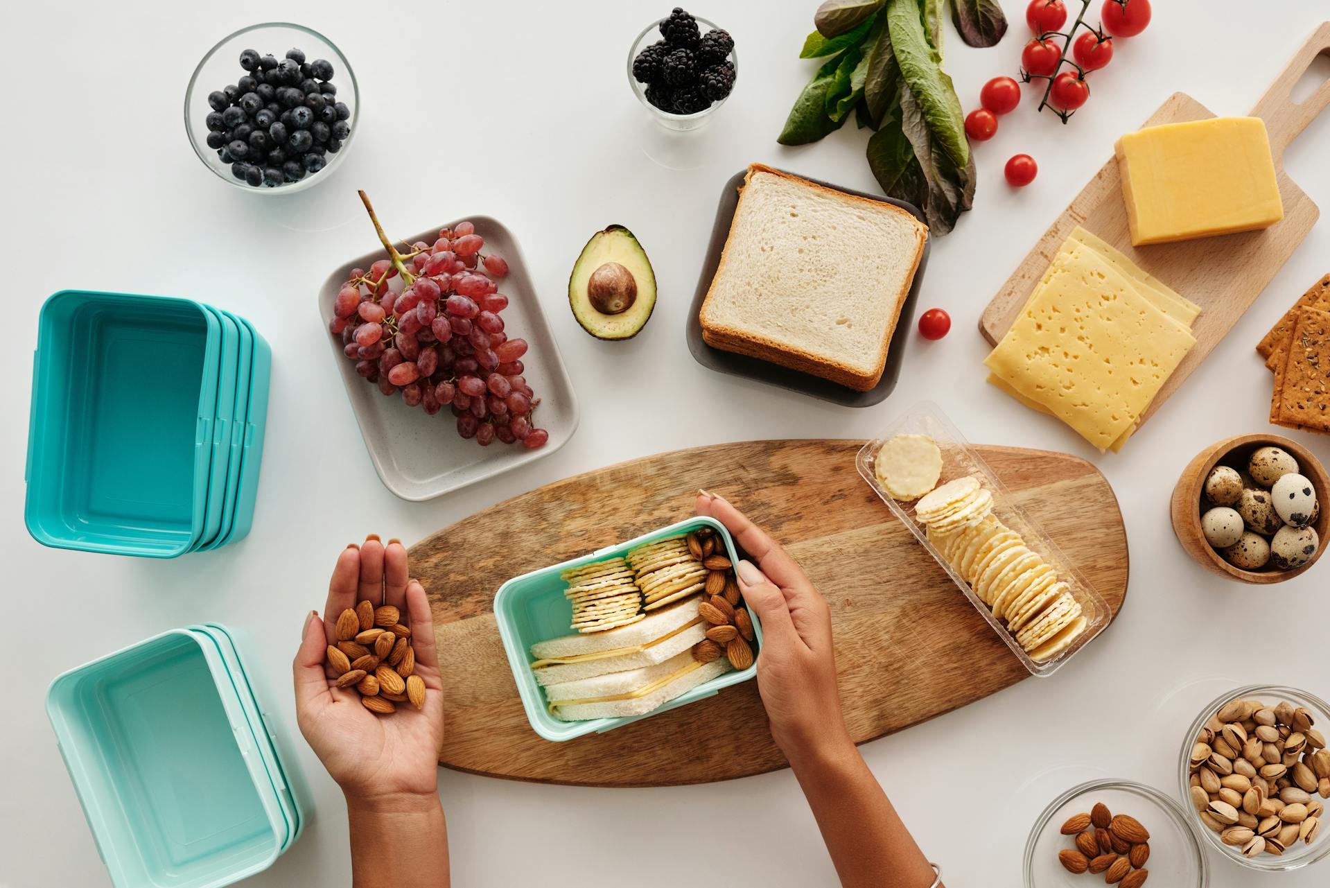 A selection of food including grapes, crackers, nuts, and cheese being put in a lunch box.