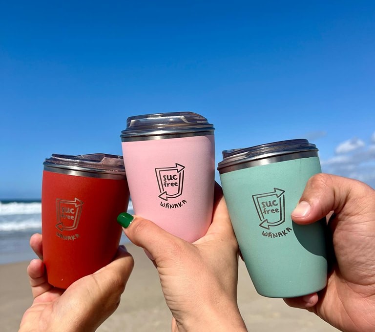 Three SUCfree cups being held together on a beach.